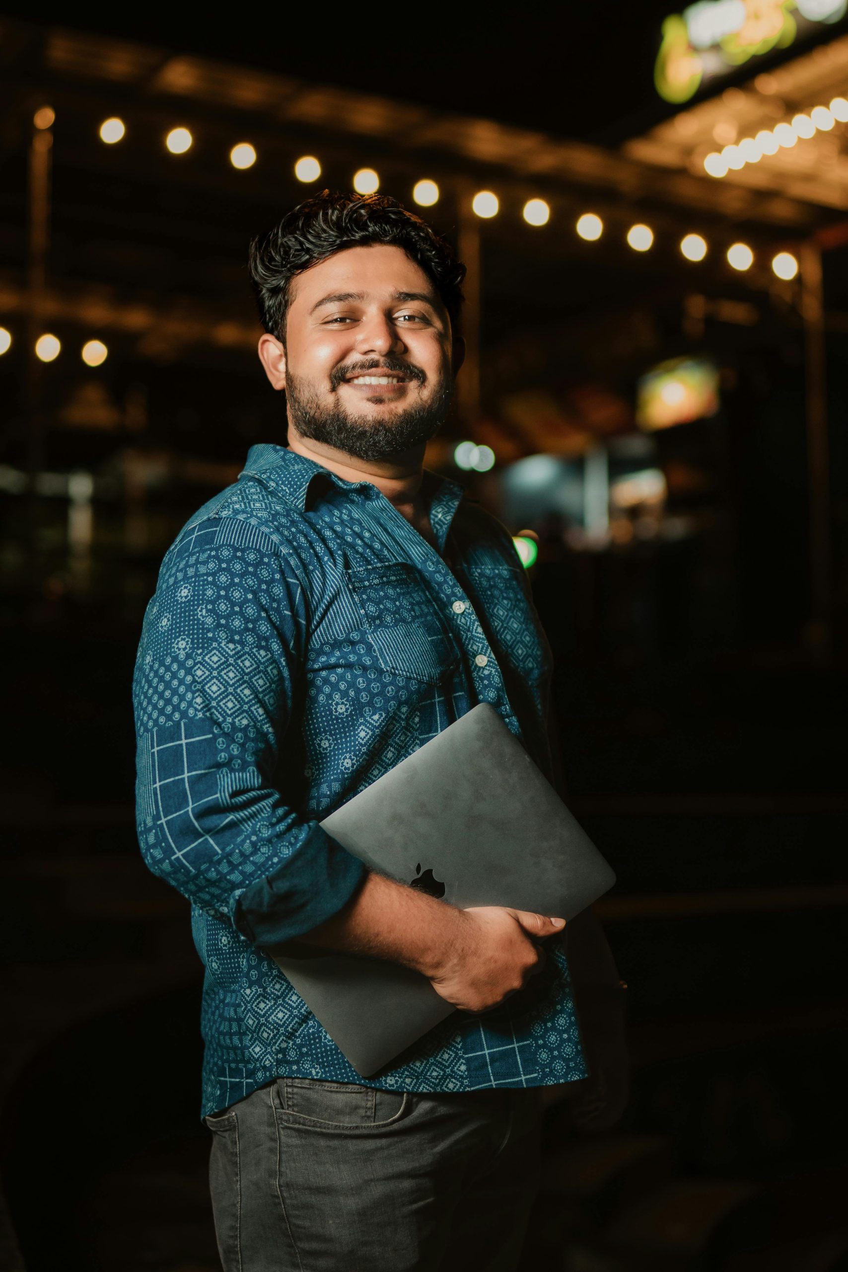 Smiling man with a beard and laptop standing outdoors at night, dressed casually in a patterned shirt.